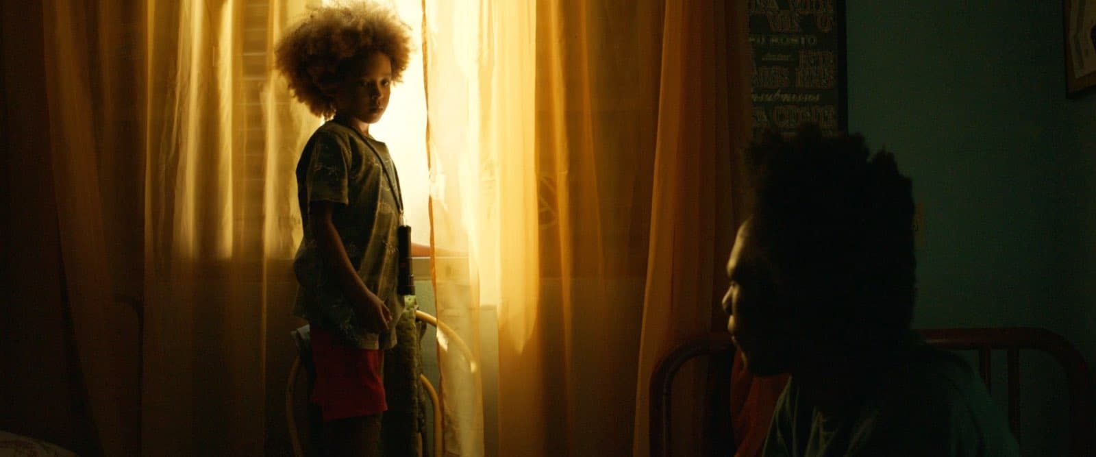 Cena de “Nosso Segredo”: uma criança em pé diante da janela, contra a luz, observada por outra pessoa no quarto