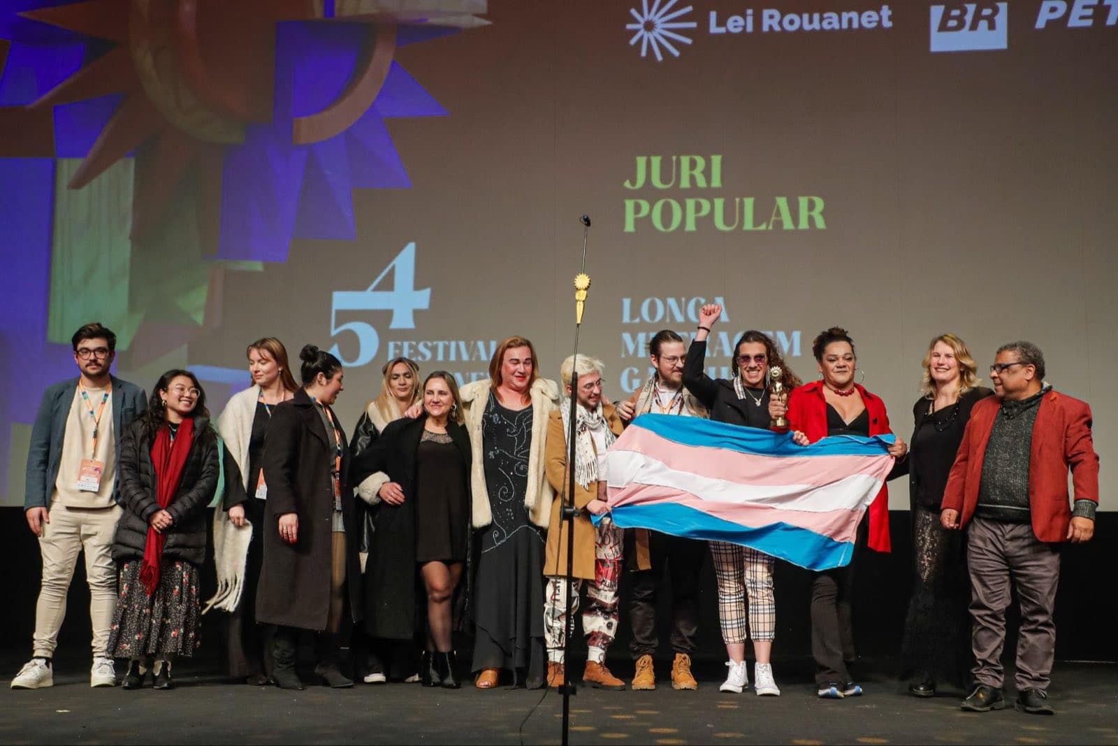 Equipe de “Filhas da Lua” no palco do Palácio dos Festivais recebendo o prêmio do Júri Popular, com a bandeira trans estendida