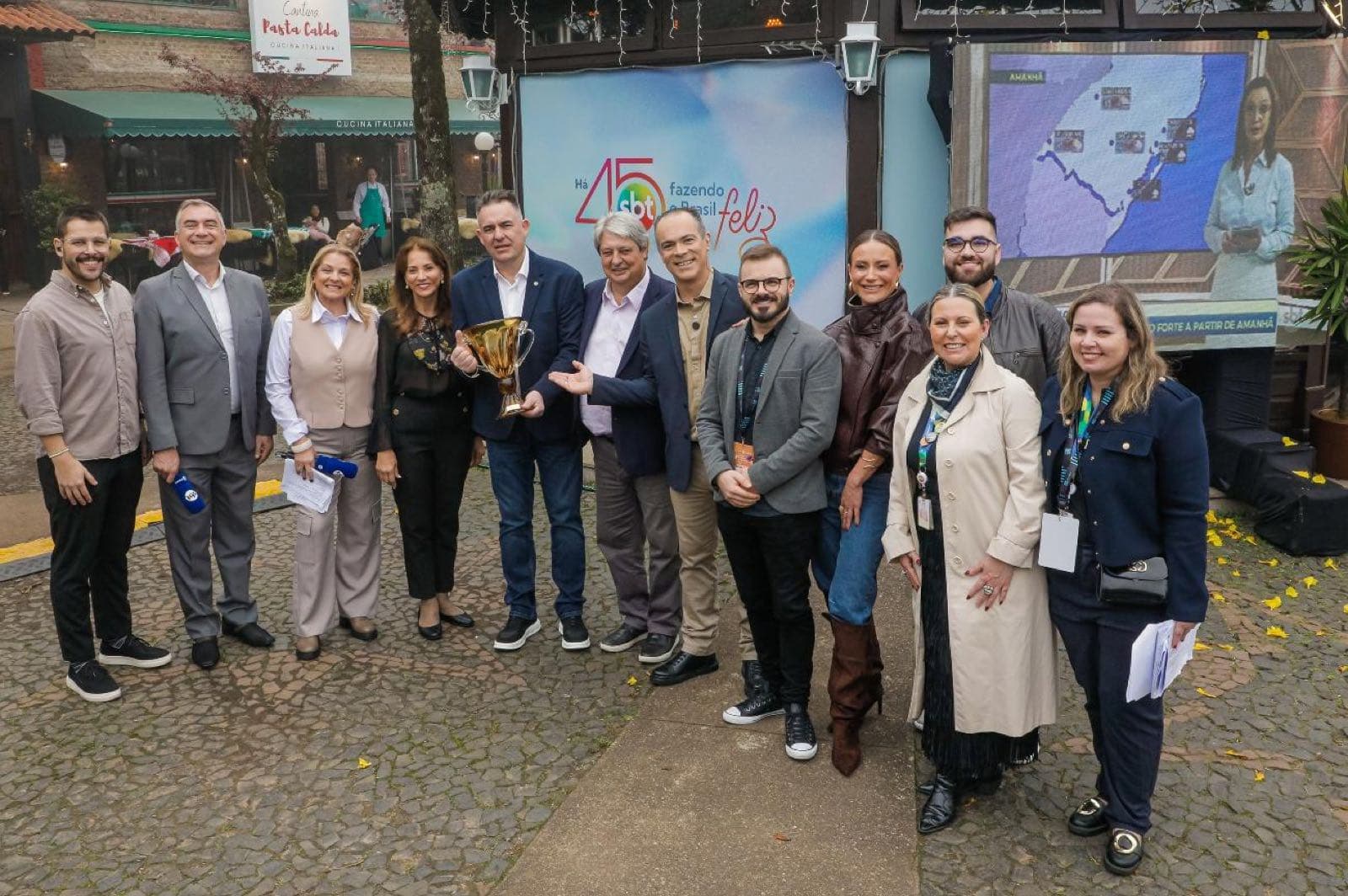 Equipe do SBT e representantes da Gramadotur reunidos com a peça da homenagem, diante do painel dos 45 anos da emissora