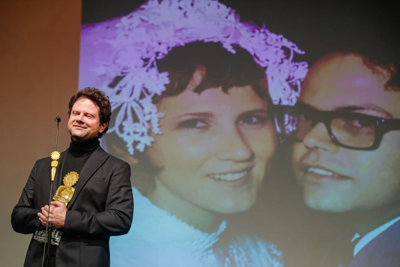 Selton Mello segura o Troféu Kikito de Cristal no palco, com a foto dos pais projetada ao fundo